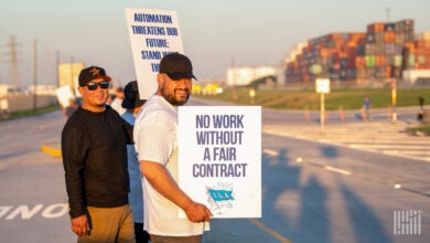 Image shows striking dockworkers holding signs with containers in background.