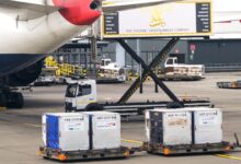 Containers pulled on carts behind the tail of a large jetliner.