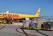 A mustard-gold DHL cargo plane at the terminal with pallets on the ground ready to load, as seen on a sunny day from the side.