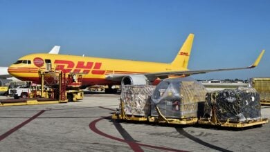 A mustard-gold DHL cargo plane at the terminal with pallets on the ground ready to load, as seen on a sunny day from the side.