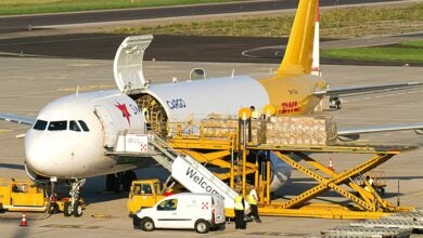 A two-tone aircraft with white paint and SmartLynx lettering, and a gold tail end with DHL logo. Cargo plane being loaded at an airport.