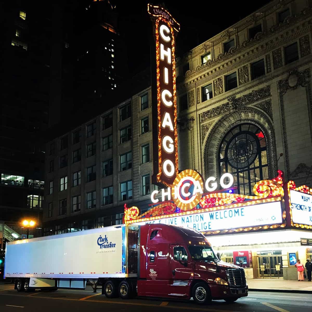 A photograph of a truck and trailer at night. The truck is in front of a brightly-lit theater with a neon marquis.