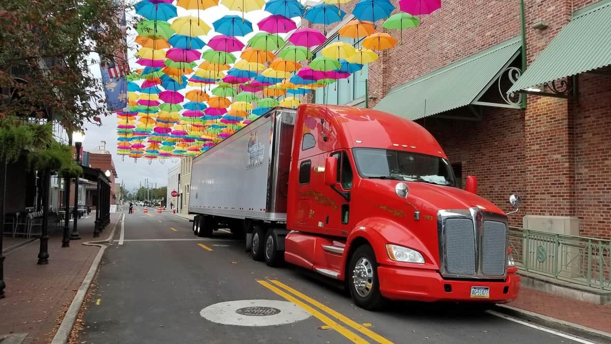 A photograph of a truck and trailer driving down a narrow street. Above the truck and the street is an art installation of rows of colorful umbrellas hanging on wires.