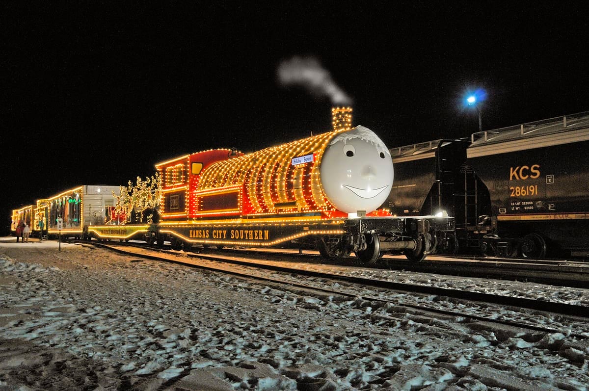 A photograph of a locomotive and railcars decorated with hundreds of holiday lights. The face of a Thomas the Train, a cartoon character, is attached to the front of a locomotive.