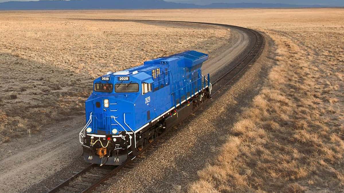 A bright blue locomotive moves down the track on a wide-open plain.