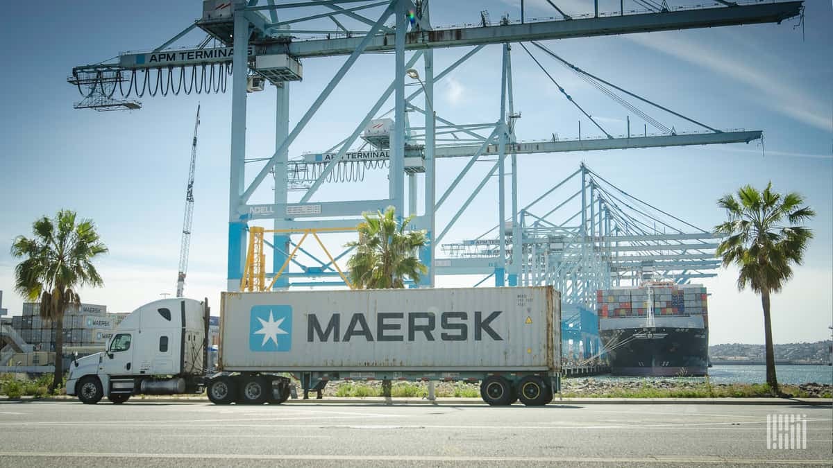 A truck carrying a Maersk container is in front of a number of cranes used to load/unload cargo ships.