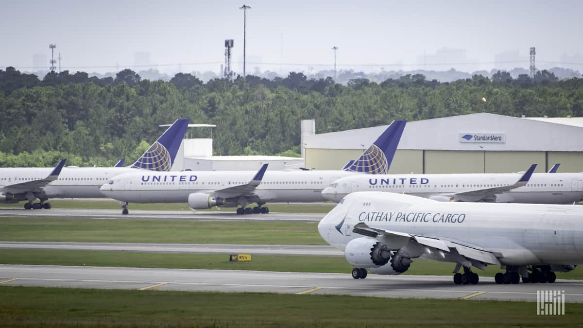 A gray Cathay Pacific 747 cargo plane takes off with white United Airlines place parked in the background.