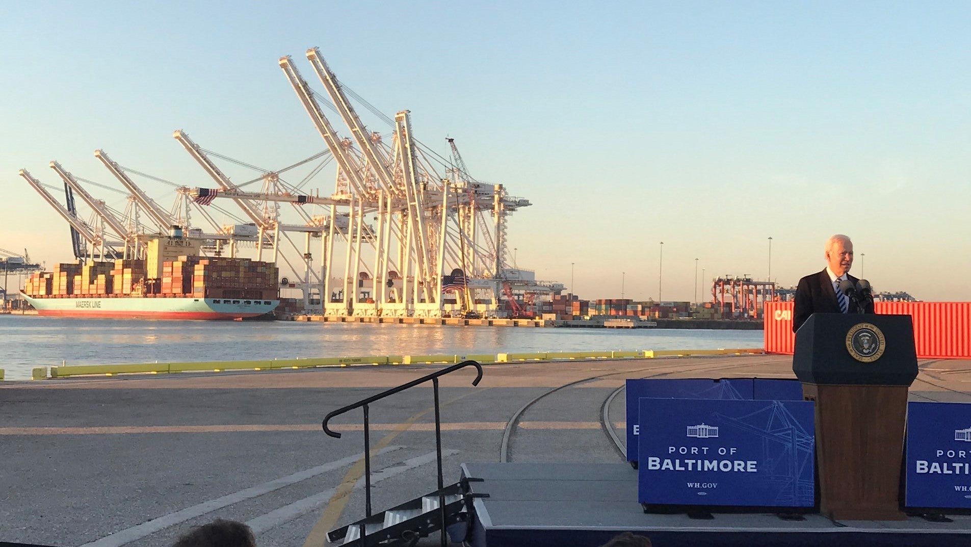 President Biden speaking at podium with port cranes and ship in the background.