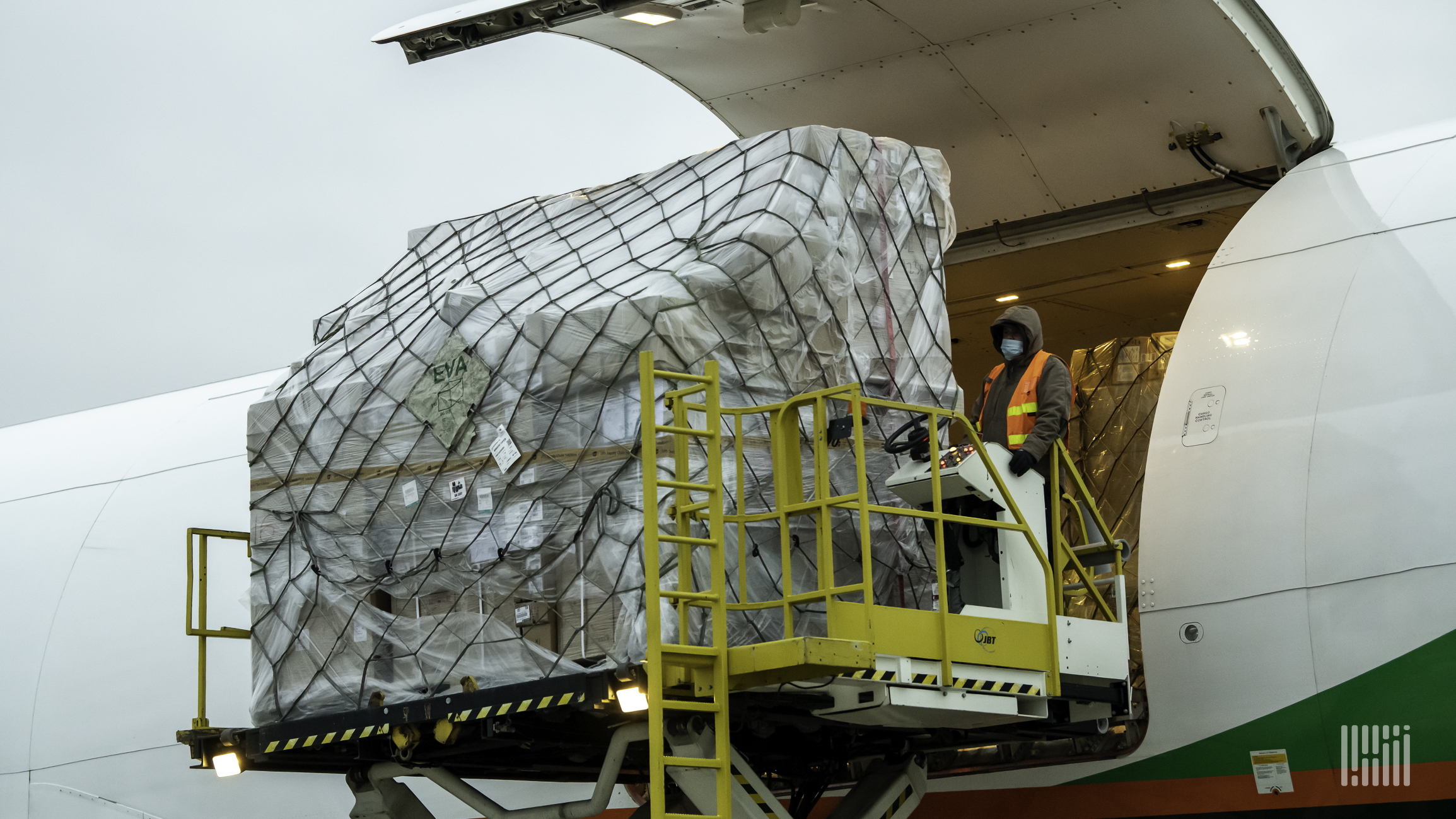 A large cargo container on a hydraulic lift being loaded into the side door of a cargo plane.