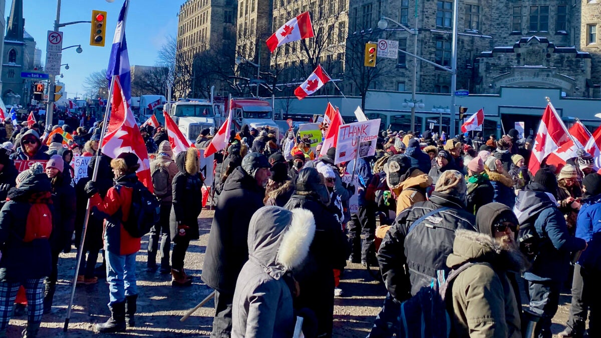 A crowd of people wave Canadian flags as trucks sit in the background.