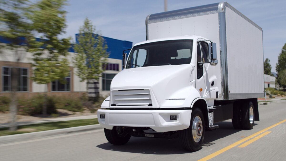 A three-quarters view of a white Xos chassis cab/box truck in motion on a street