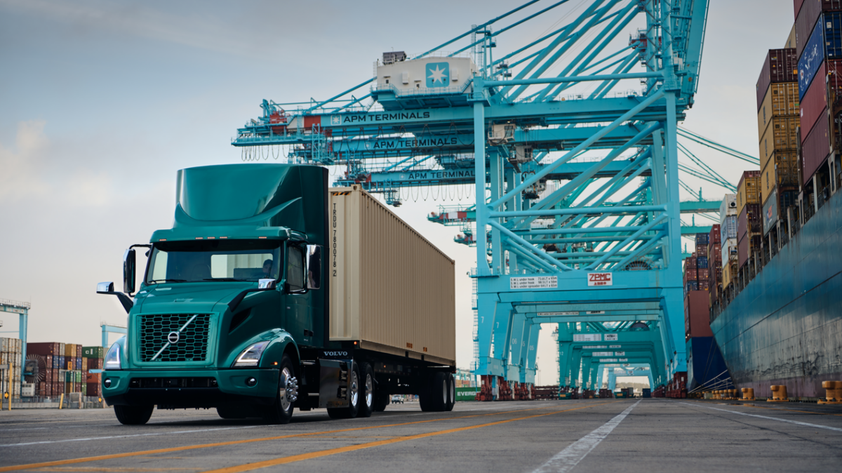 A green Volvo VNR Electric truck at the Port of Long Beach with cargo-lifting cranes and cargo containers on the right.