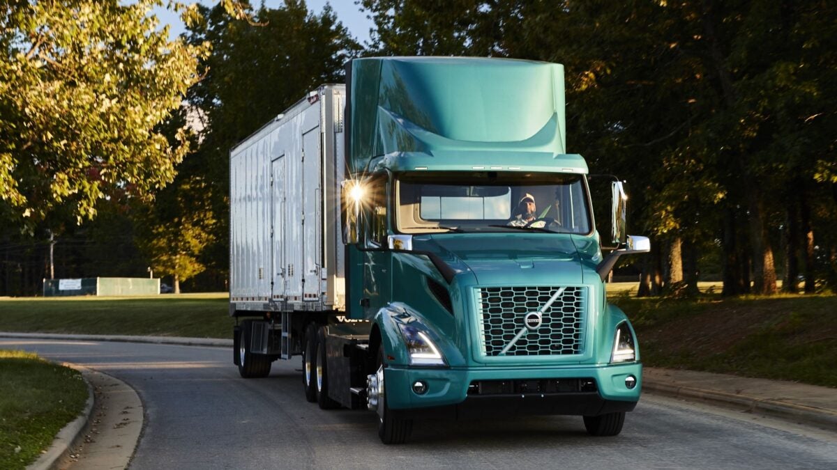 A  green Volvo VNR Electric truck driving on a tree-lined road.