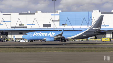 A light blue plane with Prime Air on the side rolls by a white and blue warehouse at the airport.